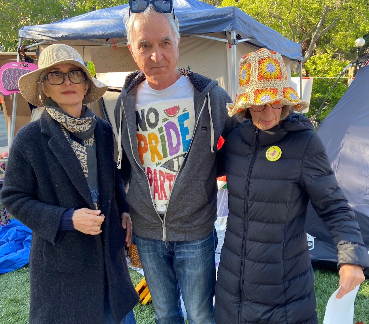 two women in funky hats and a man with a No Pride in Genocide placard tucked into his jacket